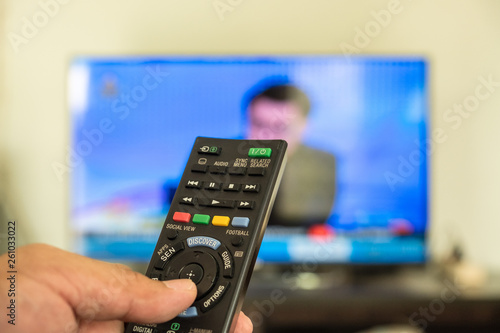 The finger is pressing on the black remote control button. TV is blurred background