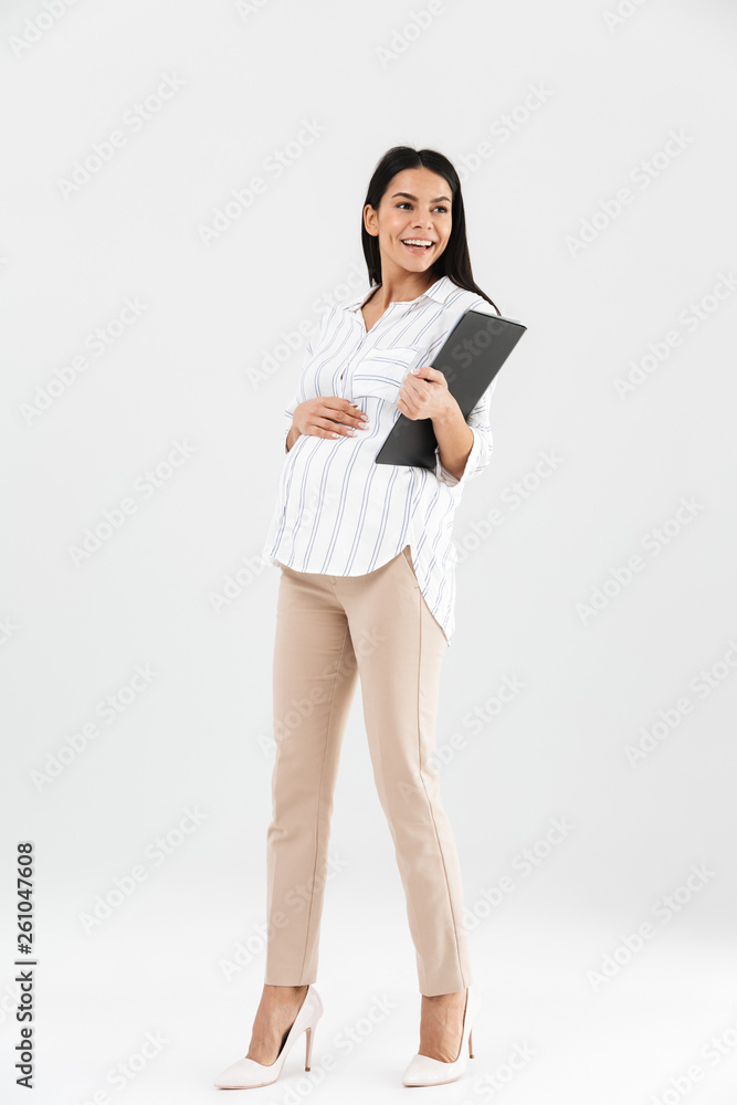 Full length photo of smiling pregnant businesswoman 30s holding clipboard and touching her big tummy Full length photo of smiling pregnant businesswoman 30s holding clipboard and touching her big tummy