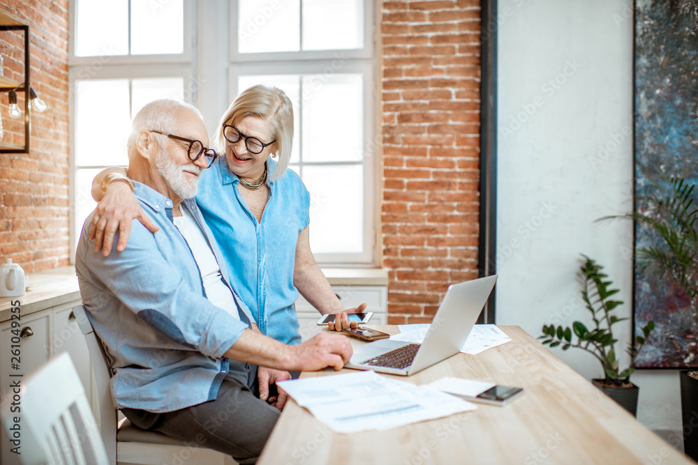 Beautiful senior couple in blue shirts hugging together working with laptop on the kitchen at home Beautiful senior couple in blue shirts hugging together working with laptop on the kitchen at home
