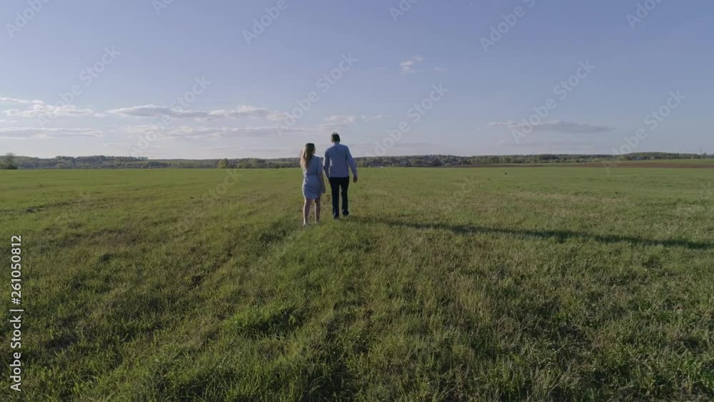 A man in a blue shirt and jeans, a girl in a blue dress, they go on the field,the girl straightens her hair man, aerial photography, the camera flies around a couple A man in a blue shirt and jeans, a girl in a blue dress, they go on the field,the girl straightens her hair man, aerial photography, the camera flies around a couple