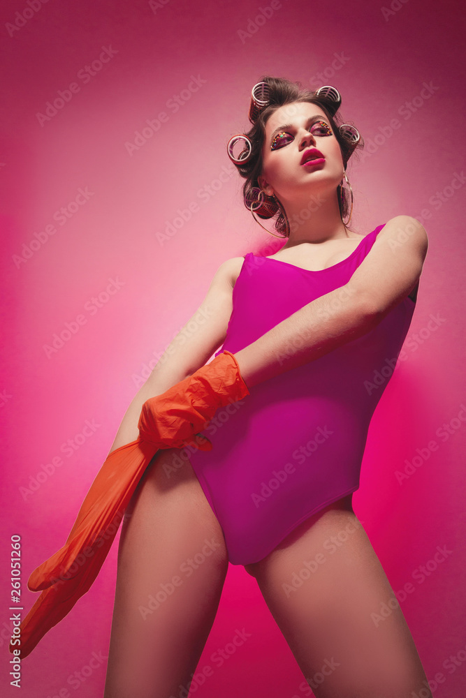Cheeky girl with latex gloves posing on pink background in body, with curlers on head. Pretty sexy woman with sweet makeup attractively posing in studio.