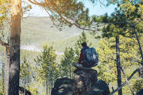 Man hiker with a large backpack sitting on a rock cliff and looking at the sprawling green valley. Freedom in travel