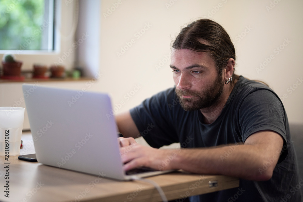 Young man with long hair using a laptop placed on a wooden desk while working from a cozy environment. Business require internet and portability most of the time Young man with long hair using a laptop placed on a wooden desk while working from a cozy environment. Business require internet and portability most of the time