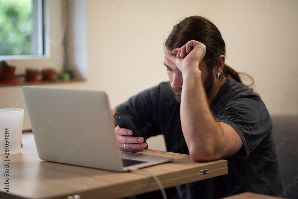 Young man with long hair using a laptop placed on a wooden desk while reading phone messages from a cozy environment. Caucasian businessman looking like a hipster Young man with long hair using a laptop placed on a wooden desk while reading phone messages from a cozy environment. Caucasian businessman looking like a hipster