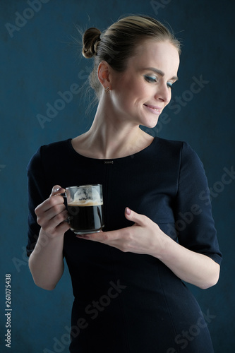 Studio portrait concept of a beautiful fashionable blonde girl standing in a business dress on a blue background drinking coffee.