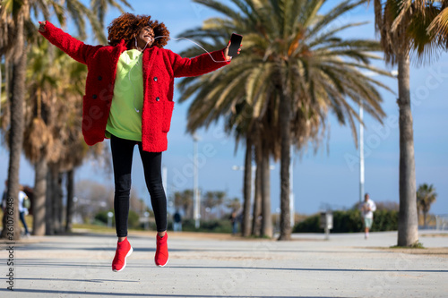 Front view of young beautiful curly african american woman jumping and dancing while listening music and smiling outdoors