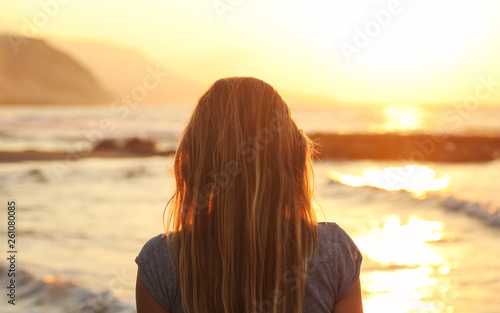 Young woman watching sunset at the beach, looking to sea, mountains in distance. View from back, only her head and hairs visible.