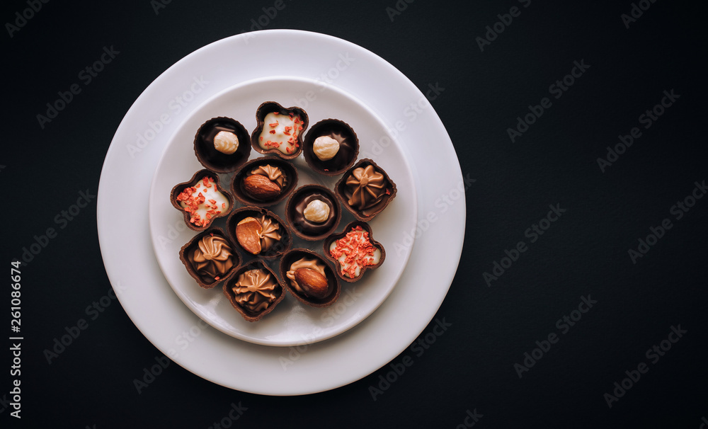 Many beautiful chocolates of different shapes and fillings lie in a white plate against the black background paper. Many beautiful chocolates of different shapes and fillings lie in a white plate against the black background paper.
