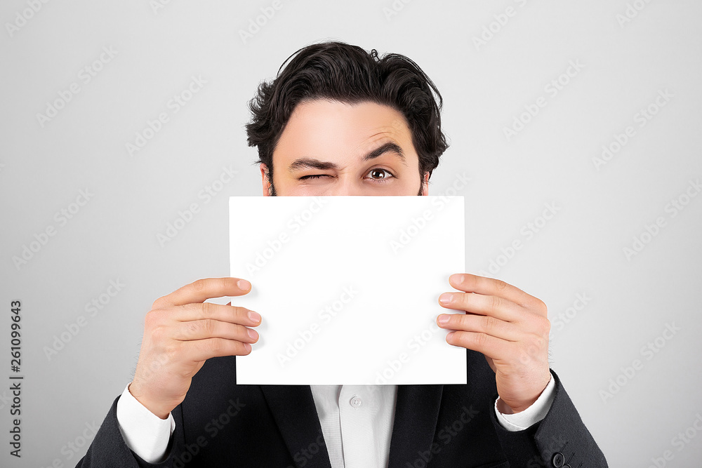 businessmen winks and shows a white sheet of paper in front of face in the camera on gray background. Area for advertising. businessmen winks and shows a white sheet of paper in front of face in the camera on gray background. Area for advertising.