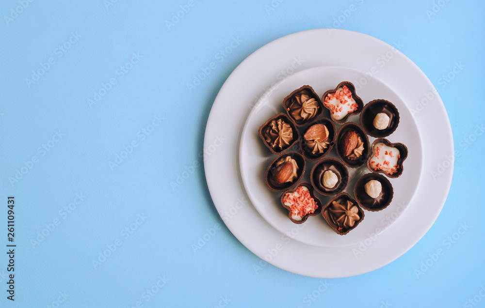 Many beautiful chocolates of different shapes and fillings lie in a white plate against the blue background paper. Copy space, top view.