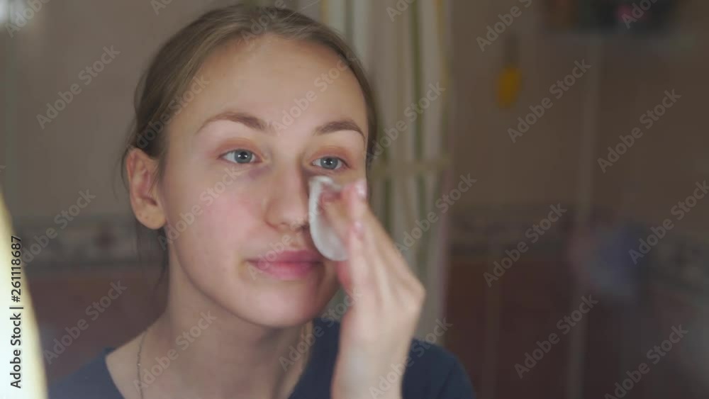 pretty young girl standing in front of a mirror in the bathroom and rubbing lotion on her face using a cotton disc pretty young girl standing in front of a mirror in the bathroom and rubbing lotion on her face using a cotton disc