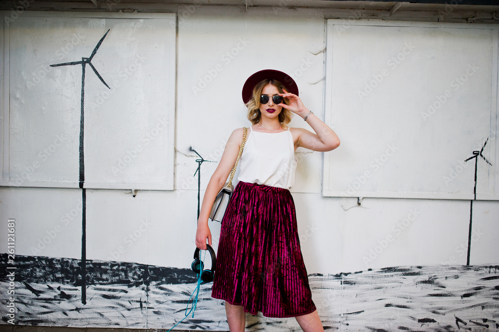 Obraz premium Fashionable and beautiful blonde model girl in stylish red velvet velour skirt, white blouse, hat and sunglasses, posed with phone and earphones.