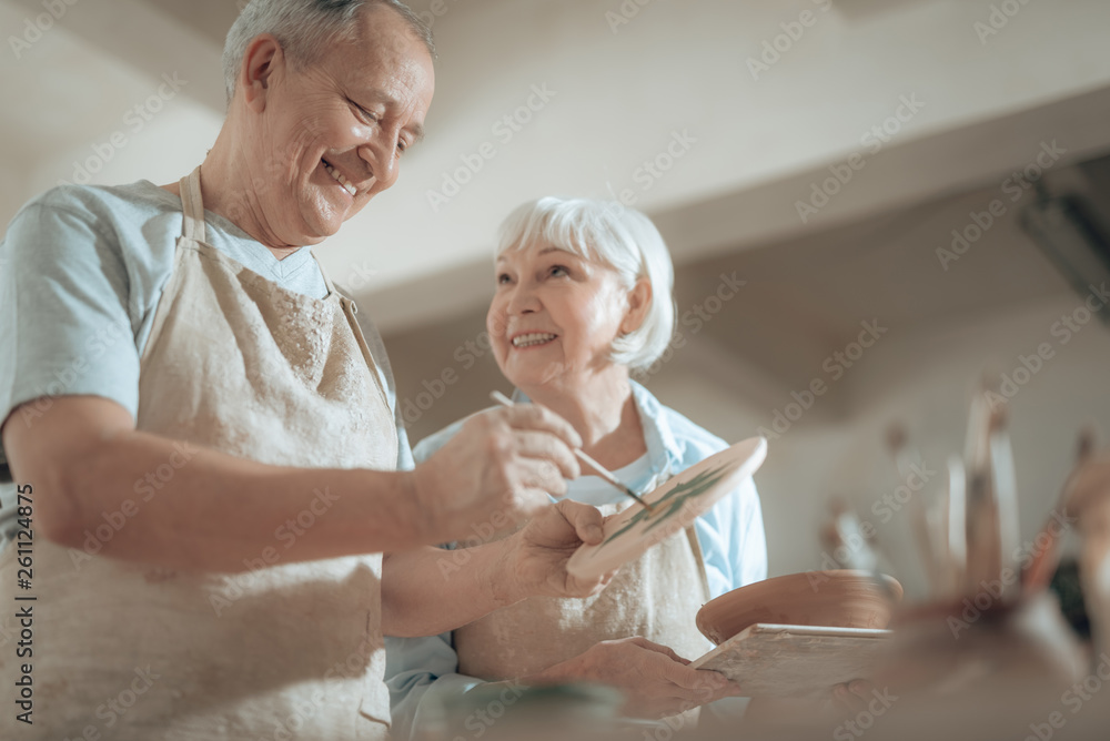 Obraz premium Low angle of smiling elderly craftsman painting decorative plate in workshop