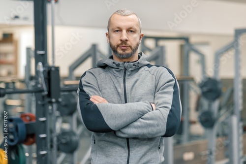 Portrait of handsome sporty man with folded hands in the gym, cross fitness instructor looking at the camera