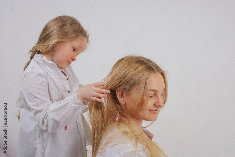 mom and daughter in white shirts with long blonde hair posing on a solid background in the Studio. a charming family takes care of each other and makes braids. mom and daughter in white shirts with long blonde hair posing on a solid background in the Studio. a charming family takes care of each other and makes braids.