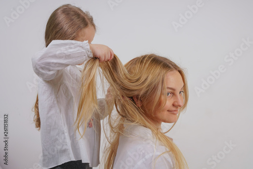mom and daughter in white shirts with long blonde hair posing on a solid background in the Studio. a charming family takes care of each other and makes braids.