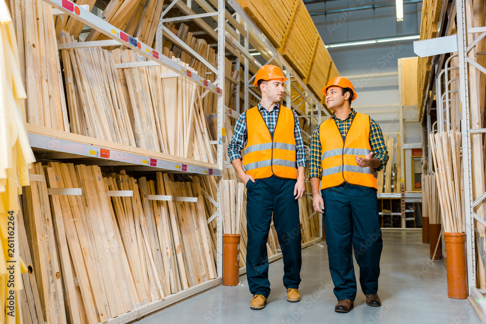 Fototapeta premium smiling multicultural workers talking while walking along racks with wooden construction materials