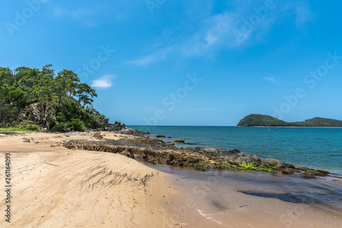Cairns, Australia - February 18, 2019: North end of warm beige tropical beach and rocks of Palm Cove with azure Coral Sea water under blue sky. Double Island in back.