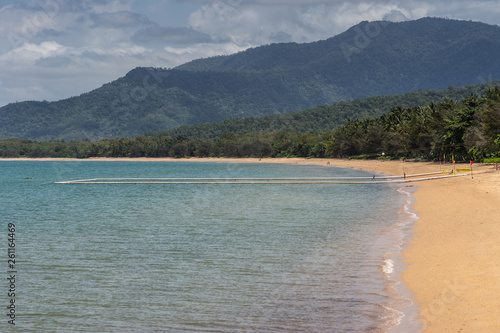 Cairns, Australia - February 18, 2019: Warm beige tropical beach of Palm Cove with azure Coral Sea water under blue cloudscape with rainforested mountains on horizon. Protected by nets and beams area 