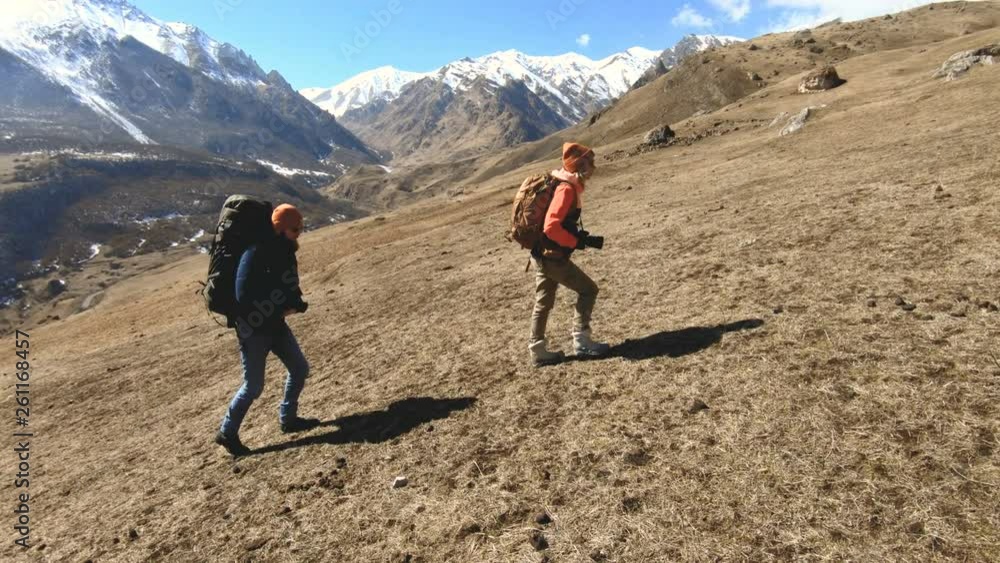 Two tourists photographers with backpacks in hats and sunglasses go up the hill on the yellow grass with cameras in their hands against the background of snowy mountains. slow motion Two tourists photographers with backpacks in hats and sunglasses go up the hill on the yellow grass with cameras in their hands against the background of snowy mountains. slow motion