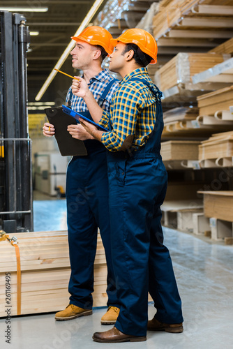 two multicultural workers in uniform holding clipboards while standing in warehouse