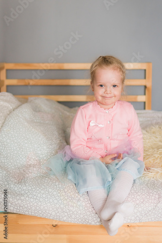 The little girl in a pink jacket sits on a bed and smiles