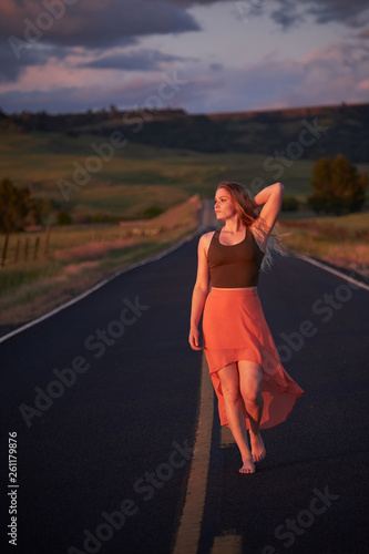 A young blonde woman in a skirt and tanktop walking barefoot down a country road at sunset.