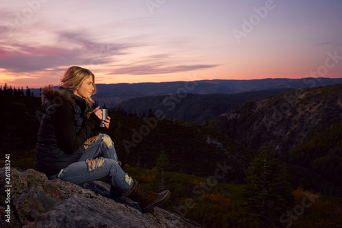 A young woman in a vest, jeans and boots drinking coffee while watching the sunset over the Sierra Nevada Mountains.