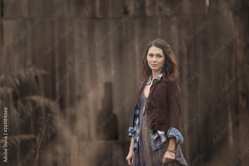 Fototapeta premium A young woman wearing a dress standing in the tall grass in a field next to a small barn on a small rural farm.