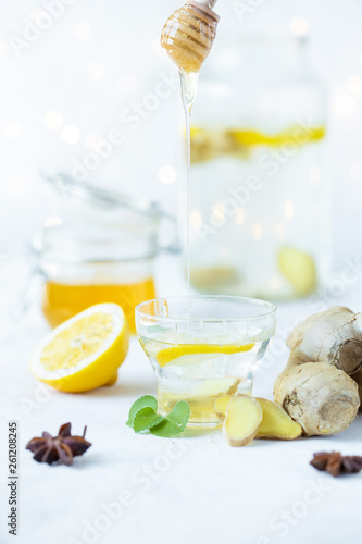 Honey is poured into a ginger drink in a cup. Ginger root, honey in a jar, lemon on a white table.