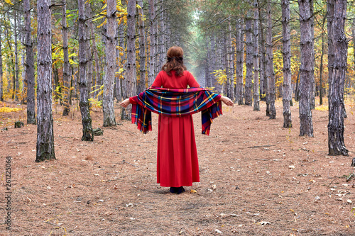 Middle-aged brunette woman in a red dress with checkered shawl posing in bright, fairy autumn pine forest. Back view