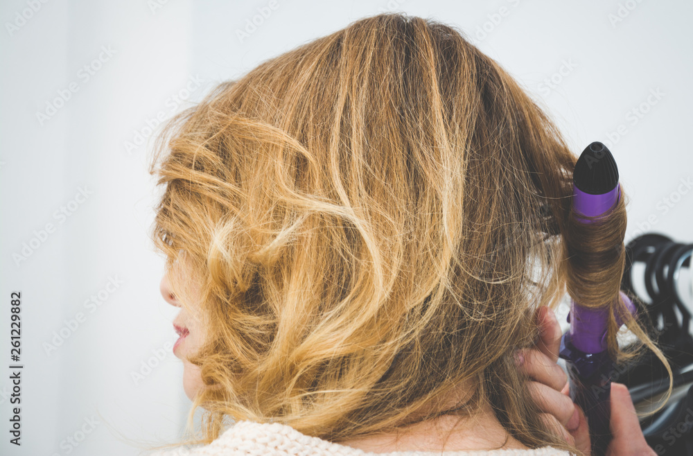 Fototapeta premium Beuty and fashion concept, Hairdresser curling woman hair with electric iron curler tong, blonde hair