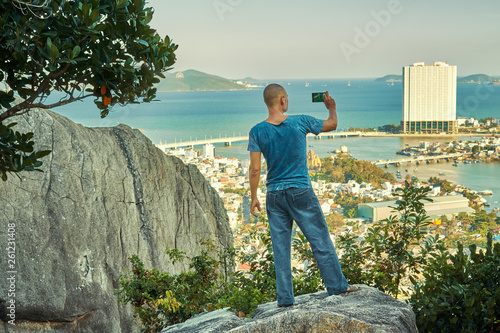 Middle aged man hiker takes selfie from the rocky mountain viewpoint overlooking coastal city at sunset time