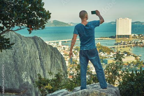 Middle aged man hiker takes selfie from the rocky mountain viewpoint overlooking coastal city at sunset time