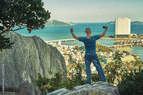 Middle aged man hiker takes selfie from the rocky mountain viewpoint overlooking coastal city at sunset time