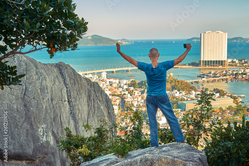Middle aged man hiker takes selfie from the rocky mountain viewpoint overlooking coastal city at sunset time