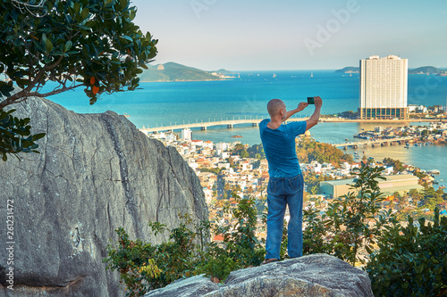 Middle aged man hiker takes selfie from the rocky mountain viewpoint overlooking coastal city at sunset time