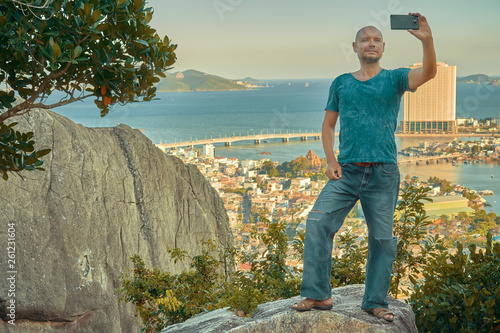 Middle aged man hiker takes selfie from the rocky mountain viewpoint overlooking coastal city at sunset time