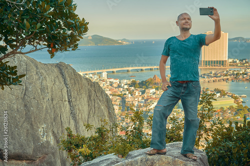 Middle aged man hiker takes selfie from the rocky mountain viewpoint overlooking coastal city at sunset time