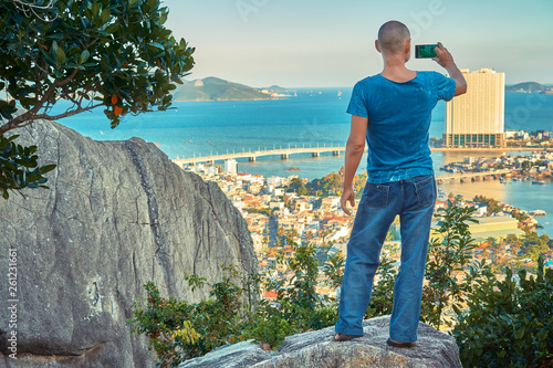 Middle aged man hiker takes selfie from the rocky mountain viewpoint overlooking coastal city at sunset time