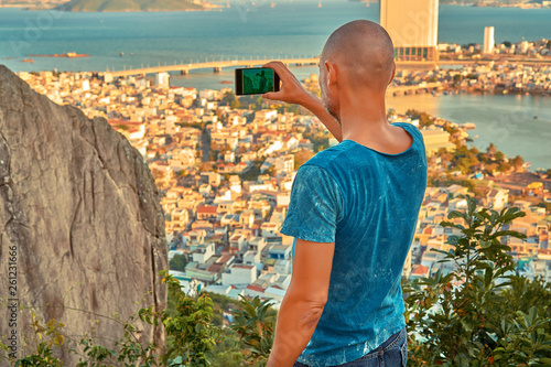 Middle aged man hiker takes selfie from the rocky mountain viewpoint overlooking coastal city at sunset time