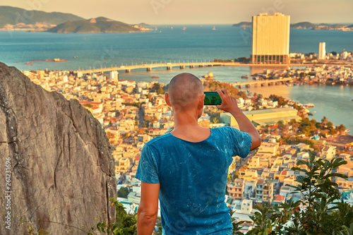 Middle aged man hiker takes selfie from the rocky mountain viewpoint overlooking coastal city at sunset time