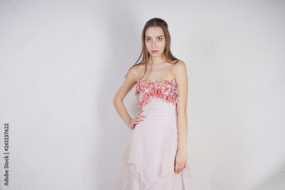 a charming young caucasian girl stands in a pink long prom dress with flower petals on her chest and poses on a white background in the Studio a charming young caucasian girl stands in a pink long prom dress with flower petals on her chest and poses on a white background in the Studio