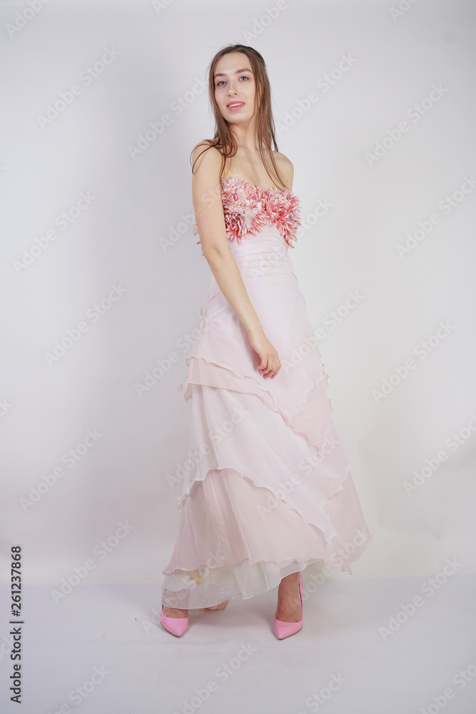 a charming young caucasian girl stands in a pink long prom dress with flower petals on her chest and poses on a white background in the Studio