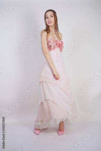 a charming young caucasian girl stands in a pink long prom dress with flower petals on her chest and poses on a white background in the Studio © goldeneden