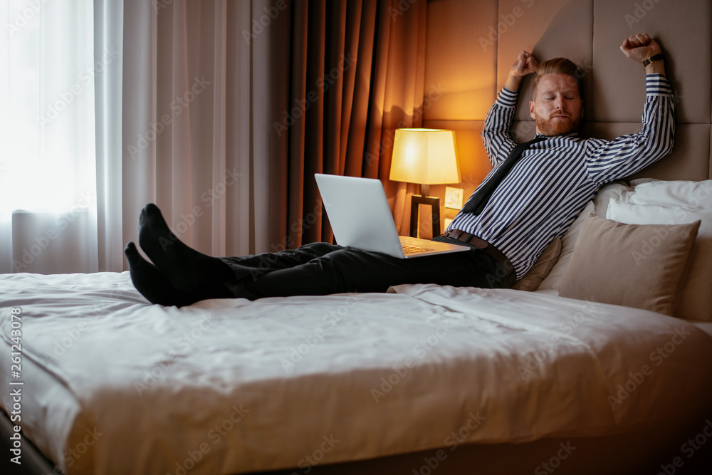Obraz premium Boss resting on a hotel bed after a long day. Manager relaxing at a hotel room with laptop beside him. Ceo enjoys his free time at a hotel.