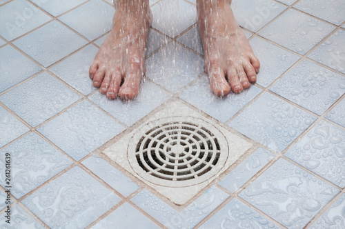 Feet standing under shower, drain on floor and  flowing water in bathroom.