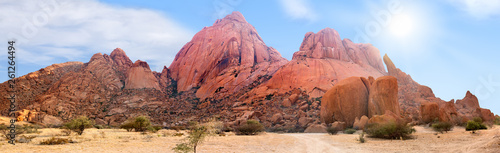 Spitzkoppe mountains range panoramic view on blue sky, clouds and bright sun background, red mountain rocks panorama landscape in Namibia, Southern Africa, beautiful tourist banner design, copy space photo