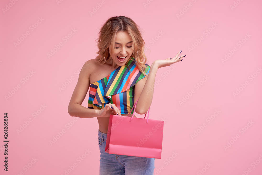Fototapeta premium Portrait of an excited beautiful smiling girl wearing colorful clothes holding shopping bag isolated over pink background. Place to sign or text on the package