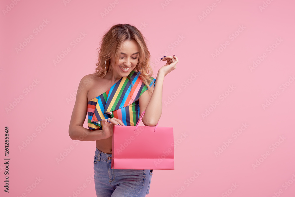 Fototapeta premium Portrait of an excited beautiful smiling girl wearing colorful clothes holding shopping bag isolated over pink background. Place to sign or text on the package